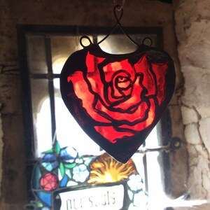 A red stained glass heart-shaped suncatcher with a rose design. The ornament is hanging in front of a window with other stained glass.