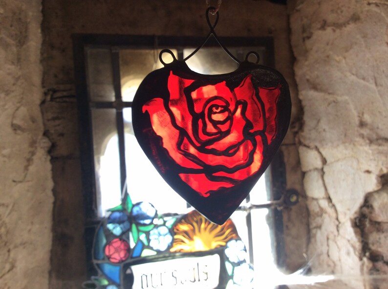 A red stained glass heart-shaped suncatcher with a rose design. The ornament is hanging in front of a window with other stained glass.