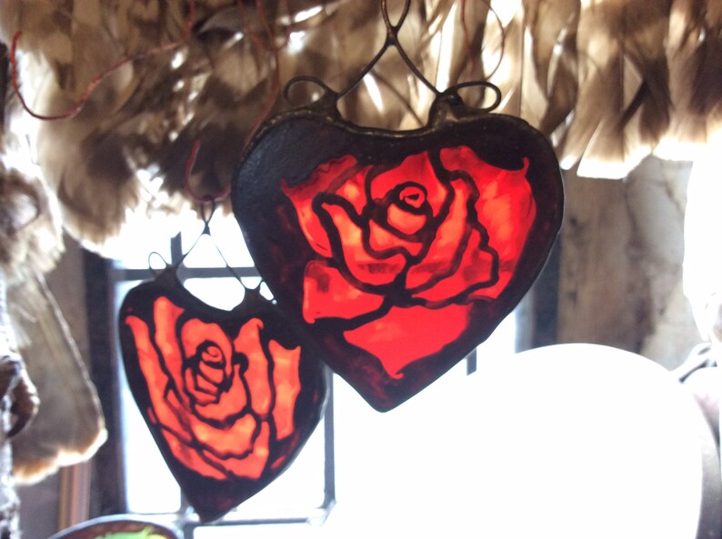 Two red stained glass hearts with black outlines, each featuring a hand painted rose design. The hearts are hanging from a hand madewire loop.