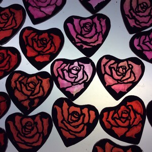 A geoup of 15 heart-shaped decorations with black outlines and red and pink rose designs. The roses are painted in a watercolour style with enamel, and then etched back before firing to sharpen the designs.