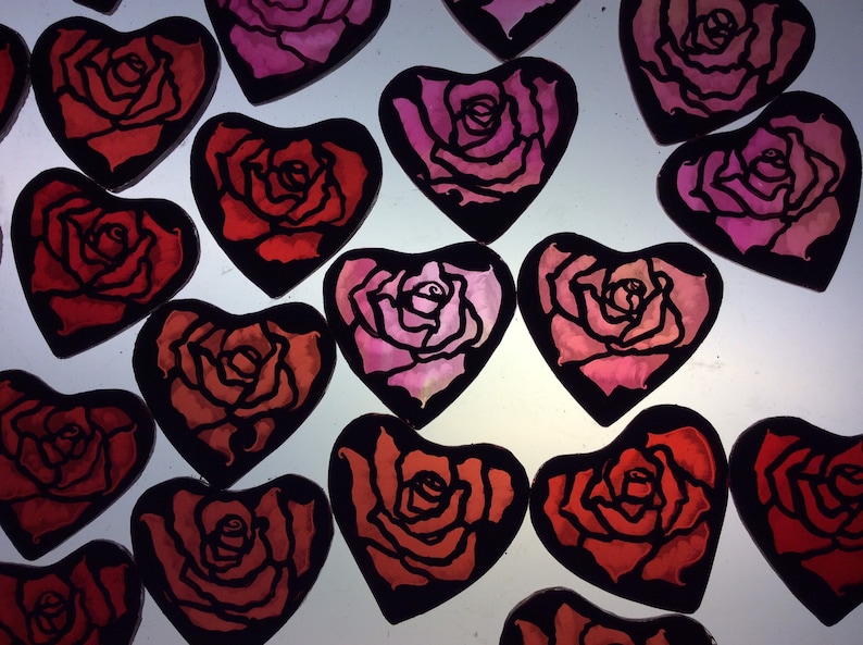 A geoup of 15 heart-shaped decorations with black outlines and red and pink rose designs. The roses are painted in a watercolour style with enamel, and then etched back before firing to sharpen the designs.