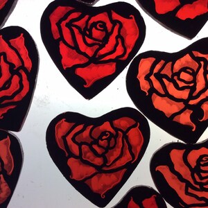 Eight red heart-shaped stained glass ornaments with black outlines, each featuring a stylised red rose design.  The roses are shown on a light box where they have been painted.  The red colour varies a little from one decor to the next.