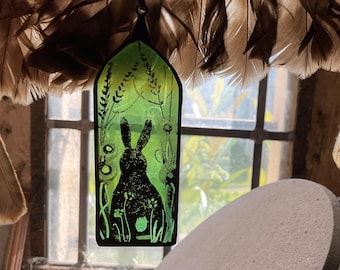 May include: A green stained glass window hanging with a black silhouette of a rabbit in a field of flowers.