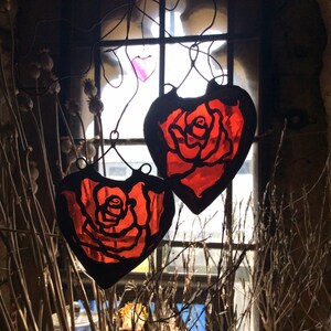 Two red stained glass heart-shaped ornaments with a rose design hanging in a window.  These sun catchers make lovely gifts for girlfriends.
