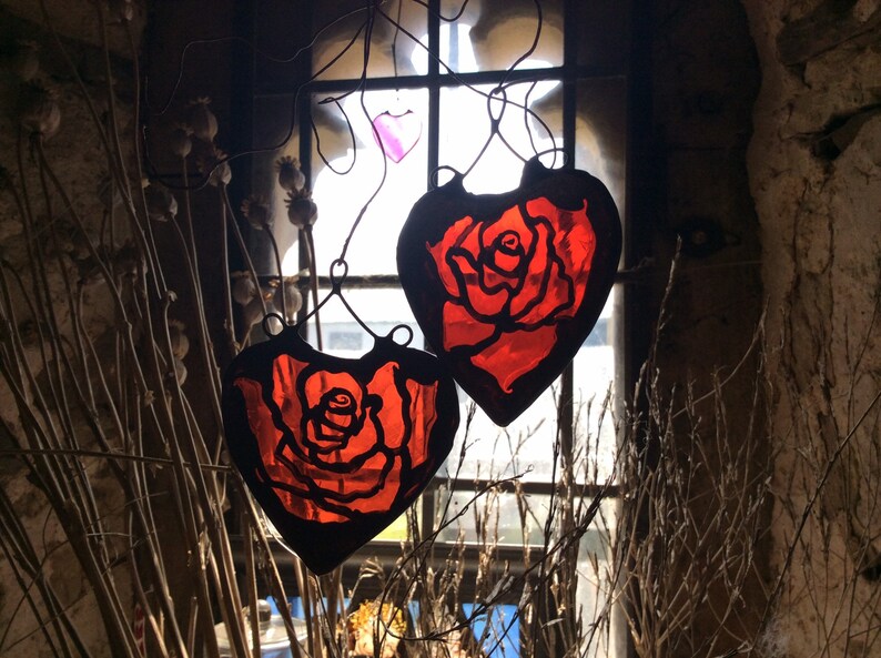 Two red stained glass heart-shaped ornaments with a rose design hanging in a window.  These sun catchers make lovely gifts for girlfriends.