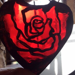 A rose design in a heart shaped piece of red stained glass.  The paint is enamel which is fired and will not fade.  The glass is surrounded by copperfoil and solder.  It is shown with daylight.