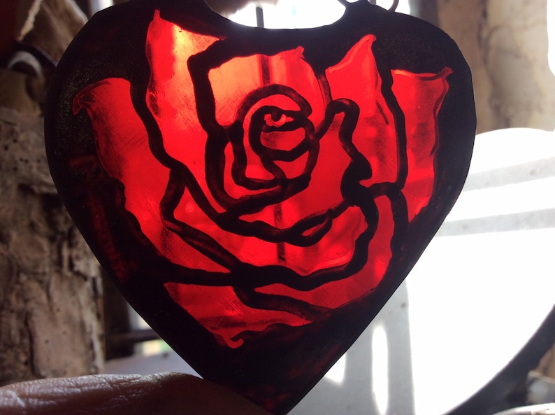 A rose design in a heart shaped piece of red stained glass.  The paint is enamel which is fired and will not fade.  The glass is surrounded by copperfoil and solder.  It is shown with daylight.