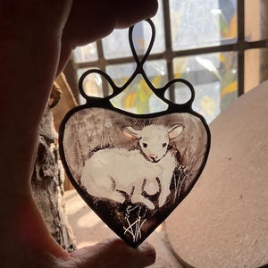 以下が含まれることがあります： 白い子羊が描かれた、ハート型のステンドグラスのオーナメント。子羊は横向きに寝そべっています。オーナメントは黒い金属製の枠で囲まれています。