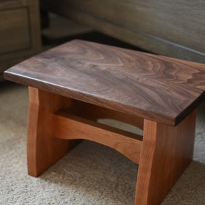May include: A wooden step stool with a walnut top and cherry wood legs. The top is smooth and has a natural wood finish. The legs are sturdy and have a slight curve at the bottom.