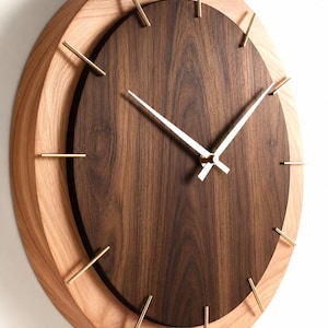 May include: A round wall clock featuring a dark brown walnut wood face set within a lighter natural wood frame. The clock has thin gold-colored metal hour markers and matching gold-colored hour and minute hands centered on the dark wood dial.