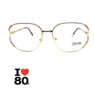 May include: Gold-toned metal spectacles with a thin, rounded frame and clear lenses. A small sticker on the right lens displays the text Steroflex. Below the glasses, a graphic features the text I, a red heart symbol, and the number 80s.