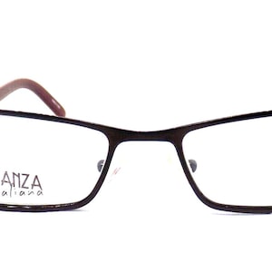 Può includere: Occhiali rettangolari con montatura in metallo marrone scuro e aste sottili. La lente sinistra presenta la scritta bianca "ELEGANZA italiana" stampata sulla superficie. Il design include un ponte e naselli regolabili per una vestibilità sicura.