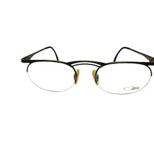 Może przedstawiać: Czarno-złote okulary z półramką z cienką, teksturowaną oprawką. Soczewki są przezroczyste, a zauszniki rozciągają się na boki. Nazwa marki jest subtelnie wyświetlana na dolnej prawej soczewce.