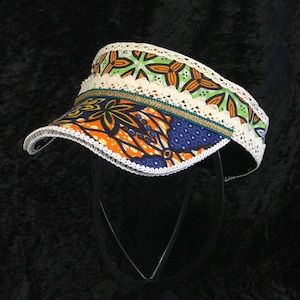 May include: A colorful visor with a white lace trim and a gold accent. The visor is made of fabric with a bright orange, green, blue, and yellow pattern.