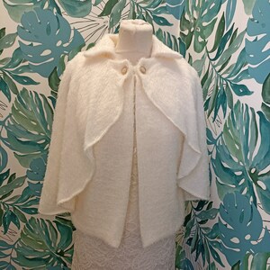 May include: White fluffy knit cape with a collar and button closure. The cape is displayed on a mannequin against a tropical leaf patterned background.