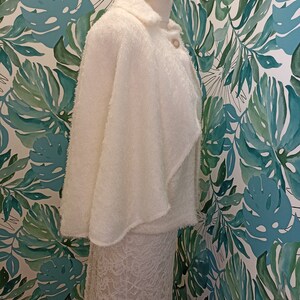 May include: A white fluffy cape with a collar and a button fastening. The cape is draped over a white lace skirt.