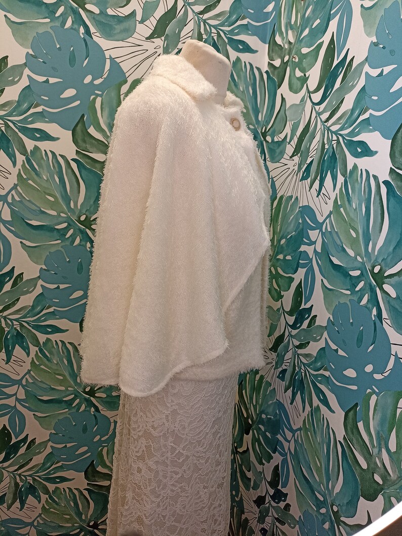 May include: A white fluffy cape with a collar and a button fastening. The cape is draped over a white lace skirt.