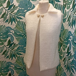 May include: White fluffy sleeveless vest with a collar and two gold buttons. The vest is on a mannequin against a background of green leaves.
