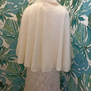 May include: A white fluffy bridal cape with a lace trim. The cape is worn over a white lace dress.