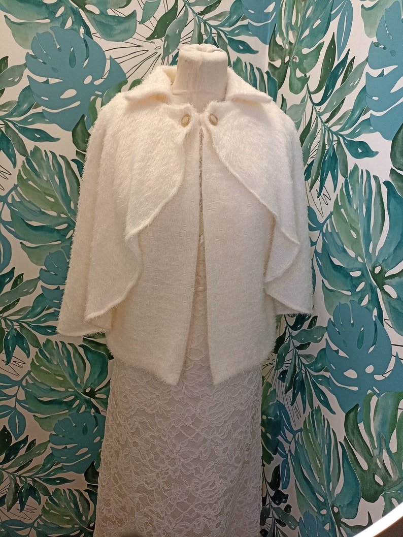 May include: White fluffy knit cape with a collar and button closure. The cape is displayed on a mannequin against a tropical leaf patterned background.