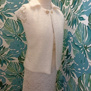 May include: White fluffy sleeveless cardigan with a collar and a gold button fastening. The cardigan is displayed on a white mannequin against a green and white floral background.