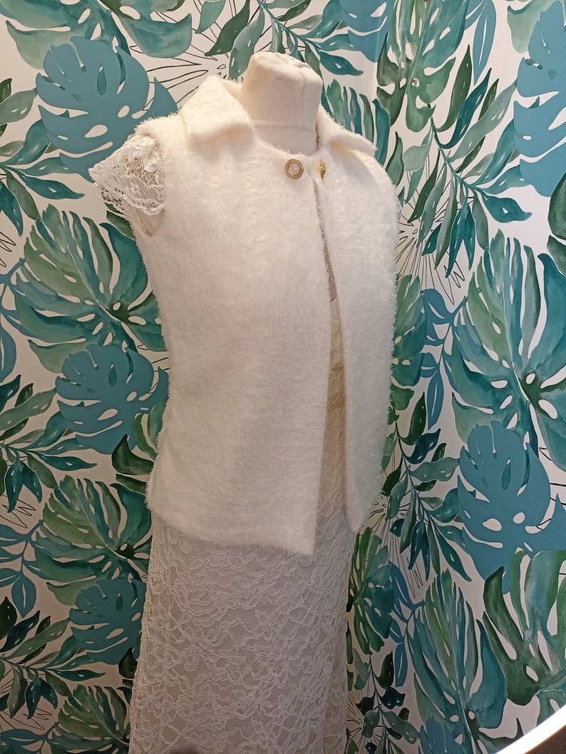 May include: White fluffy sleeveless cardigan with a collar and a gold button fastening. The cardigan is displayed on a white mannequin against a green and white floral background.