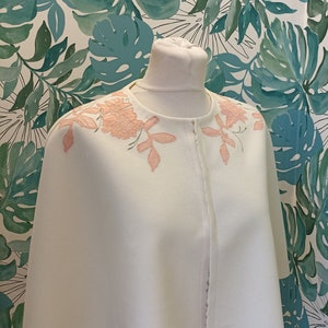 May include: A white cape with a delicate pink floral embroidery design. The cape has a simple, elegant style and is perfect for adding a touch of sophistication to any outfit.