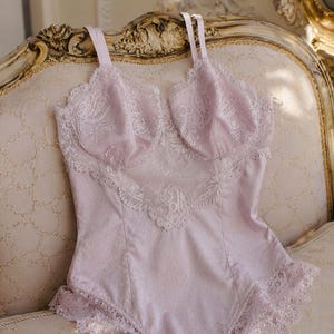 May include: A light purple bodysuit with lace detailing. The lingerie features a sweetheart neckline and delicate lace trim along the edges. The fabric has a subtle polka dot pattern. The straps are thin and adjustable.
