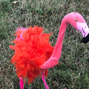 Puede incluir: Títere de flamenco de peluche con un cuello largo y curvado y un cuerpo rosa brillante. El flamenco tiene un pico negro, un ojo amarillo y un pecho de plumas naranjas esponjosas. El títere está suspendido por cuerdas negras sobre un fondo de hierba verde.