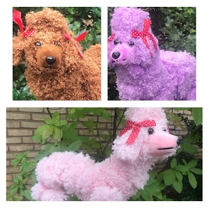 May include: Three plush poodle dog decorations in brown, purple, and pink. Each poodle has curly fur, a black nose, and a red bow with white polka dots. The dogs are set against a background of green foliage.