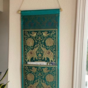 May include: Teal fabric wall organizer with two pockets, featuring an embroidered design of peacocks and elephants. The organizer has a gold trim and fringe.