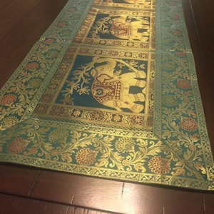 Può includere: Un runner da tavolo color turchese con un bordo dorato e un motivo ripetuto di elefanti in oro e marrone. Gli elefanti sono circondati da motivi floreali.