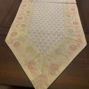 May include: Cream-colored table runner with a floral pattern and gold trim. The runner has a repeating pattern of small, stylized flowers in the center and larger floral designs along the edges. The trim features gold and peach-colored accents.