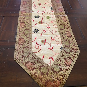 May include: A white table runner with red and green floral embroidery. The runner has a brown, gold, and red border with a floral pattern.