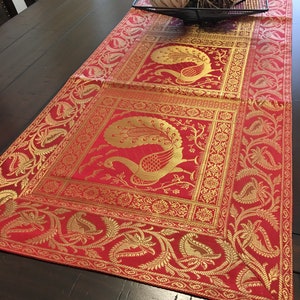 May include: Red table runner with gold embroidery featuring peacocks and floral designs. The runner is on a wooden table with decorative items in the background.