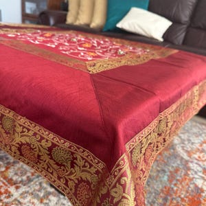 May include: A deep red tablecloth with a gold floral border. The tablecloth is covering a table, with a sofa and cushions visible in the background. The gold border features detailed floral designs.