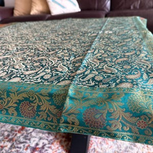 May include: A teal table runner with a paisley pattern and gold accents. The runner has a teal border with a floral design.