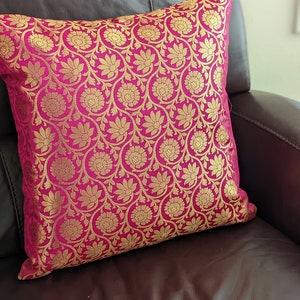 May include: A pink throw pillow with a gold floral pattern. The pillow is sitting on a brown leather couch.