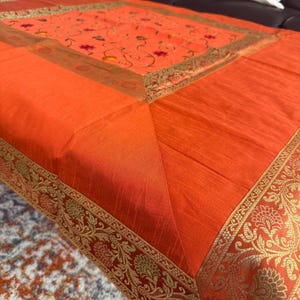 May include: An orange tablecloth with a detailed floral design in the center and a gold border. The tablecloth has a square shape and is made of a shiny fabric. The gold border features an intricate pattern.
