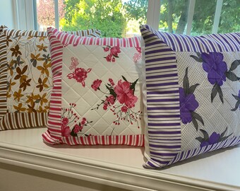 Puede incluir: Tres cojines decorativos con estampados florales y a rayas. Uno presenta diseños florales marrones y beige, otro tiene flores rosas sobre un fondo blanco acolchado, y el tercero muestra flores moradas.