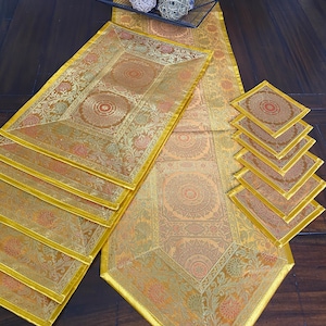 May include: A table setting featuring a gold and orange patterned table runner, placemats, and coasters. The runner and placemats have a detailed floral design. Decorative spheres in a black wire basket sit at the top.