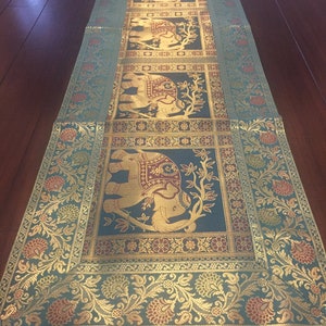 Könnte beinhalten: Ein türkisfarbener Tischläufer mit goldener Bordüre und einem sich wiederholenden Muster aus Elefanten und floralen Designs. Die Elefanten sind golden und haben mahagonifarbene Akzente.