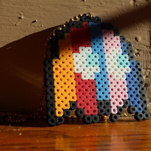 May include: A colourful beaded necklace with a Pac-Man shaped charm featuring the transgender pride flag colours: light blue, pink, and white, with a blue and white stripe on the top and a red and yellow stripe on the bottom.
