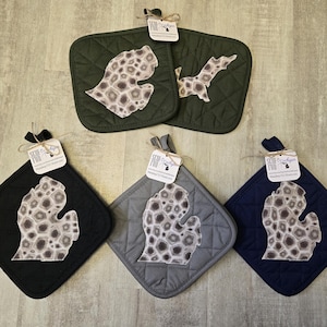 May include: Five quilted pot holders in various colors: black, gray, navy, and olive green. Each pot holder features a Michigan state outline with a unique pattern. The pot holders have a diamond pattern and a loop for hanging.