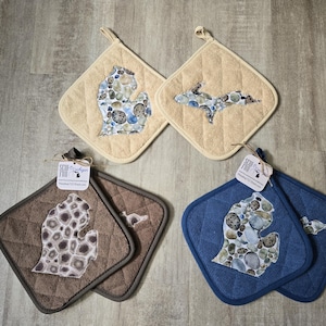 Puede incluir: Cuatro agarraderas acolchadas en beige, marrón y azul. Cada una presenta un contorno bordado del estado de Michigan, relleno con un patrón de piedras. Las agarraderas tienen un lazo para colgar y una etiqueta que dice "Sew Fun Michigan".