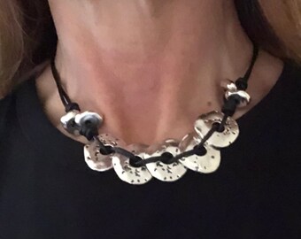 May include: A silver necklace with a black leather cord and multiple round silver pendants. The pendants have a textured surface and are connected by the cord.