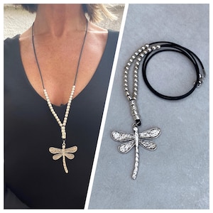 Può includere: Una collana con un ciondolo a forma di libellula argentata, un cordoncino in pelle nera e perline argentate. La libellula ha una superficie testurizzata ed è un simbolo popolare di trasformazione e cambiamento.