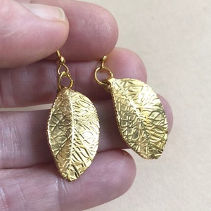 Puede incluir: Un par de pendientes de hoja de oro con una superficie texturizada. Los pendientes cuelgan y tienen un peque&ntilde;o bucle en la parte superior.