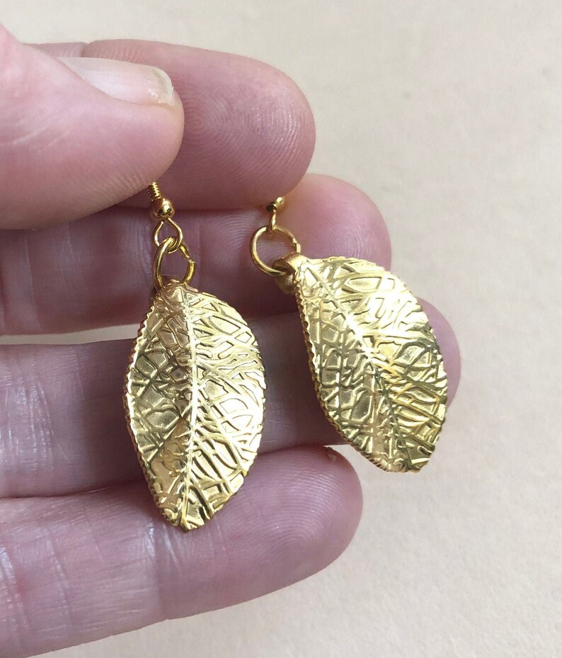 Puede incluir: Un par de pendientes de hoja de oro con una superficie texturizada. Los pendientes cuelgan y tienen un peque&ntilde;o bucle en la parte superior.