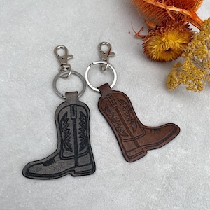 Könnte beinhalten: Zwei Leder-Schlüsselanhänger in Form von Cowboy-Stiefeln. Einer ist dunkelgrau, der andere braun. Beide haben silberfarbene Schlüsselringe und Verschlüsse. Die Stiefel haben detaillierte Nähte und sind vor einem hellgrauen Hintergrund mit getrockneten Blumen platziert.